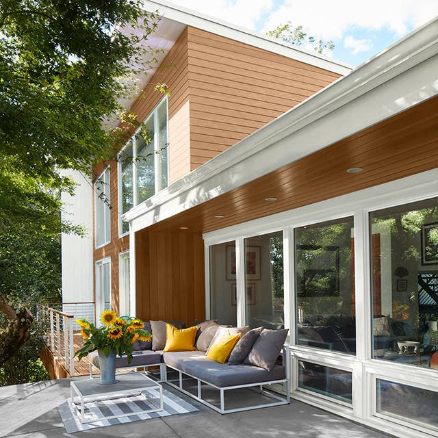 A gray deck with a blue outdoor couch with yellow pillows and a white-framed glass table with yellow flowers backs up to a one-storey enclosed porch with brown wood siding and white trim, attached to a two-storey home with light brown siding and white trim.