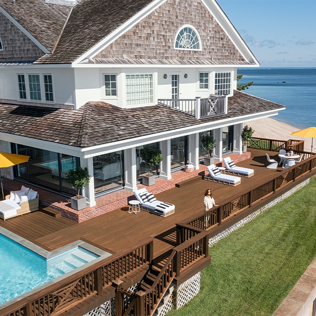 A lovely waterside home with white-painted siding and columns, taupe stained shingles, and a brown wrap around deck with lounge chairs, yellow umbrellas, and a pool.  
