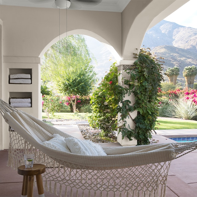 An inviting covered pool patio with white-painted stucco walls, climbing vines, and a hammock overlooking flowering red foliage and a scenic mountain range.