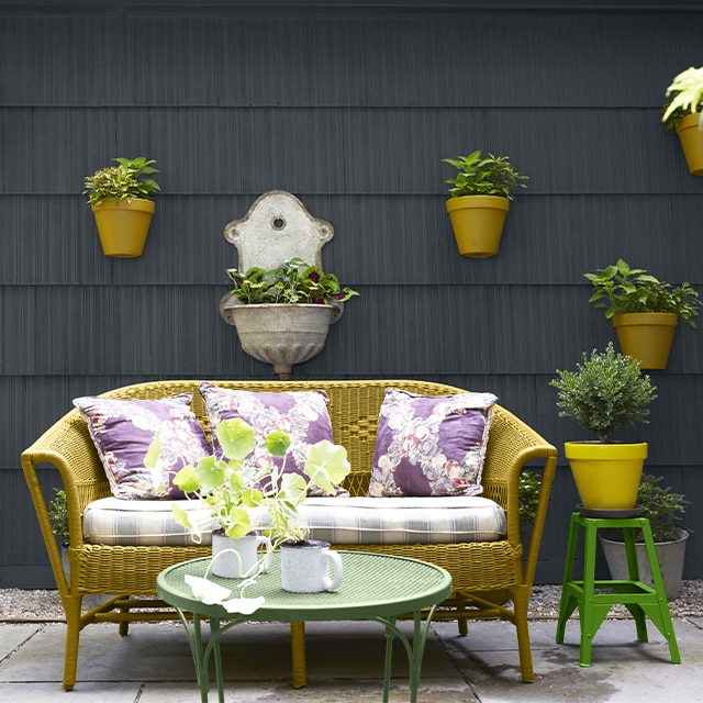 A relaxing patio space features a cushioned yellow-green wicker settee with colorful pillows and two green tables in front of light black-painted siding with attached hanging potted plants.