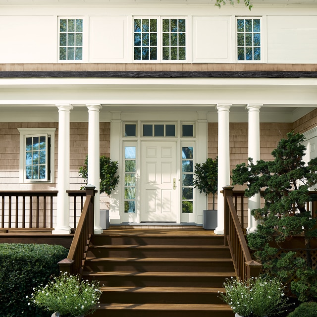 This lovely home features white-painted siding, taupe stained shingles and a brown staircase leading to a front porch with brown railings, white columns and a white front door.