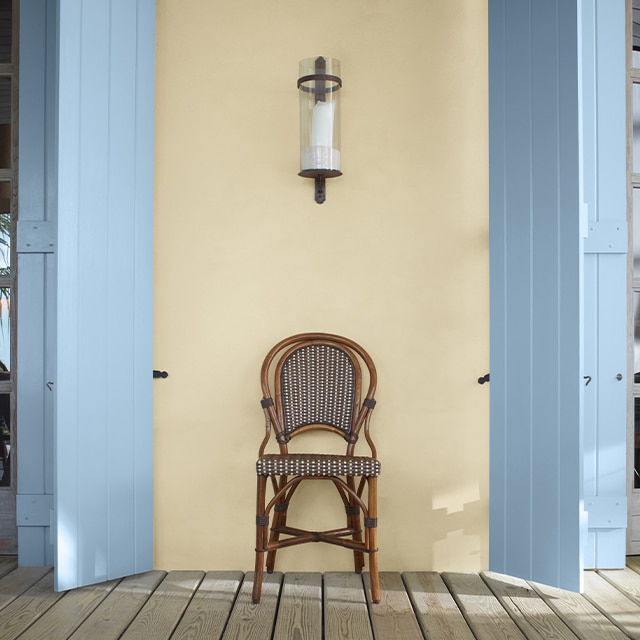 A porch featuring a chair between two light blue painted shutters against a pale yellow-painted wall.