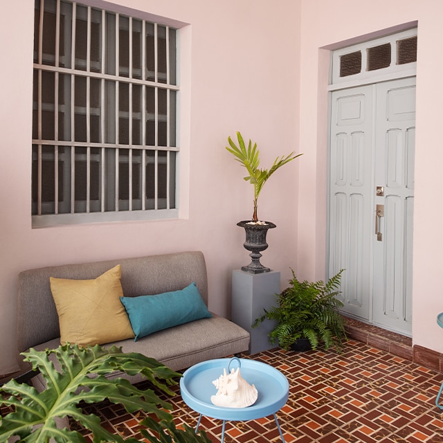A pretty porch sitting area with pink-painted walls and a gray door features a small sofa, blue table, tropical plants, and a colorful red tile floor. 