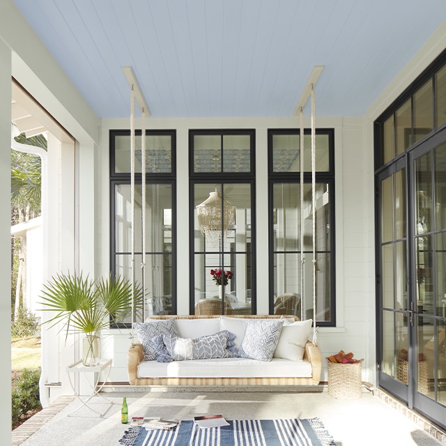 A white porch with window trim painted black, a white floor, blue and white area rug, and a hanging swing.