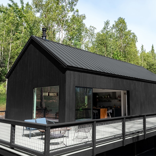 A modern style one-story home with black stained siding, large windows, and a spacious wrap around gray porch with black railings.