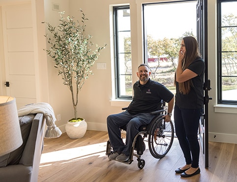 A man in a wheelchair and a woman in a home with beige-painted walls, large houseplant and wood floors.