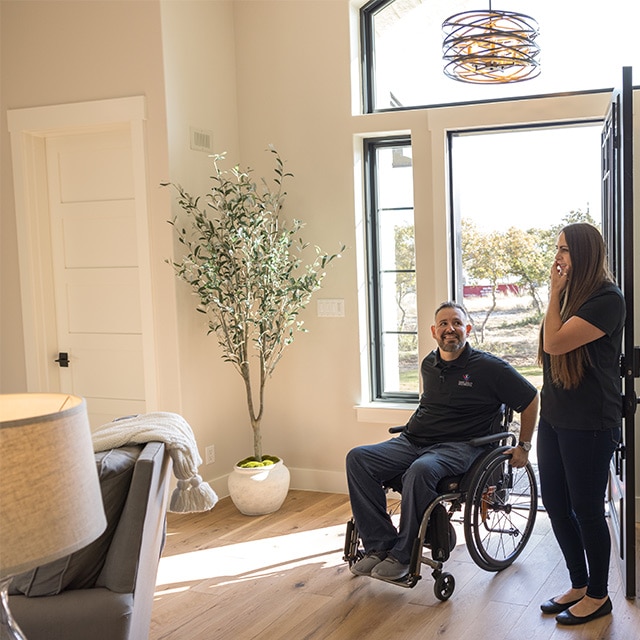 A man in a wheelchair and a woman in a home with beige-painted walls, large houseplant and wood floors.