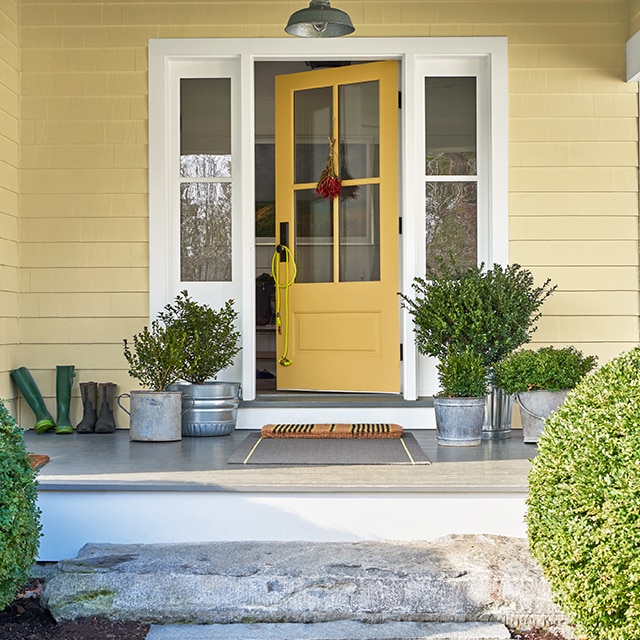 The front porch of a yellow-painted house with a yellow door, white trim, plants in metal pails, and two round shrubs out front.