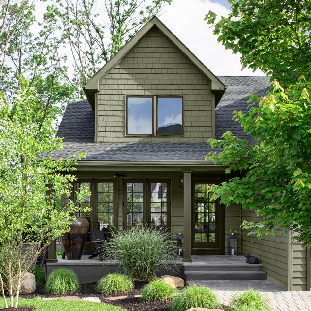 Jolie maison verte avec porche et marches en pierre entourée d’un luxuriant écrin de végétation.