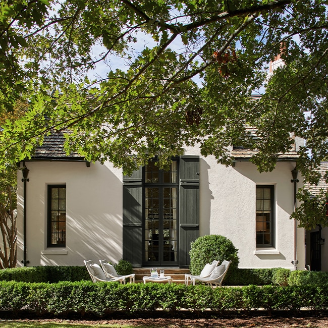 A creamy white-painted stucco home with tall centre glass doors and dark green door shutters and trim, and steps to patio furniture surrounded by a green hedge and trees.