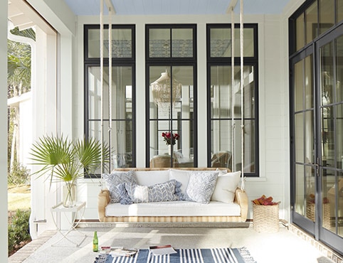 A white-painted covered porch with black-trim windows and door, a porch swing and blue and white stripe rug.