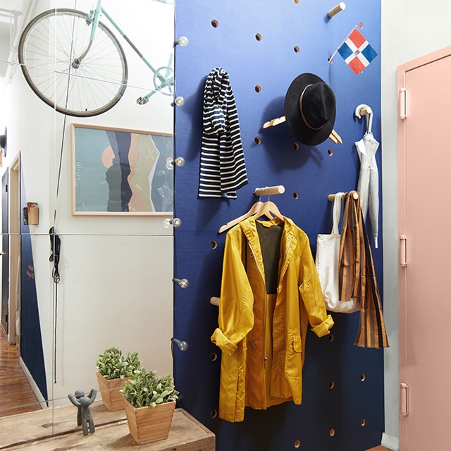 A loft entryway with a dark indigo blue pegboard wall holding hats, scarves, a jacket and umbrellas with a light pink front door.