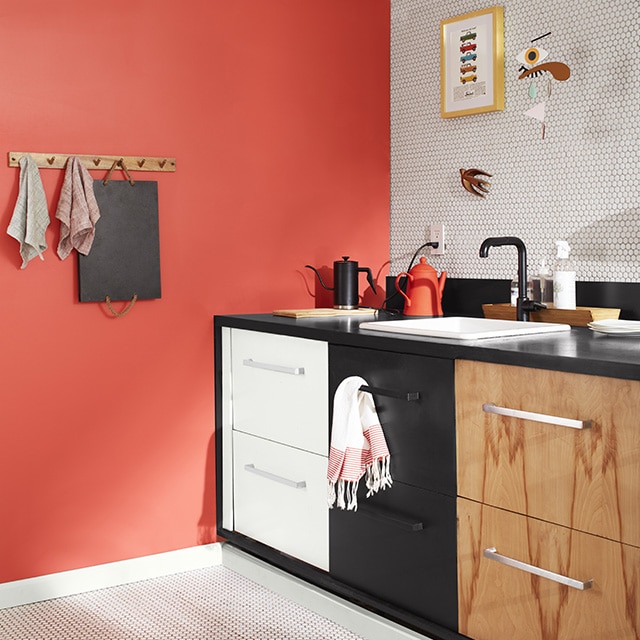 A sleek kitchen corner with a coral painted wall, a white tiled backsplash wall and floor, white upper wall and exposed pipes, and three sets of drawers: one painted white, the second black, and the third woodgrain. 