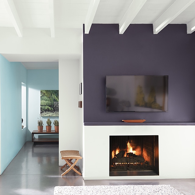 A modern white living room space with a rich, purple-painted accent wall and mantel over an active fireplace, and pretty blue hallway and back walls.
