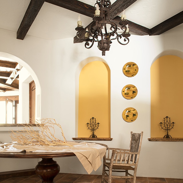 Vaste salle à manger blanche avec deux alcôves jaunes, des assiettes décoratives jaunes, des poutres en bois foncé, un lustre métallique de style rustique et une table ronde avec une chaise.