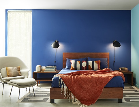A bedroom with two blue-painted accent walls, a wooden bed frame, rust coloured throw, contemporary beige chair, and reading lamps on both sides of bed.