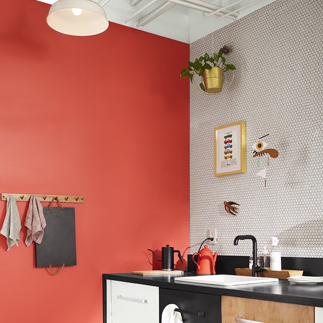 A sleek kitchen corner with a coral painted wall, a white tiled backsplash wall and floor, white upper wall and exposed pipes, and three sets of drawers: one painted white, the second black, and the third woodgrain. 