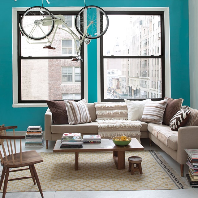 A bright, city apartment living room with a turquoise-painted accent wall, white side wall and trim, two large windows, beige and brown furnishings, and a bike hanging from exposed pipes.