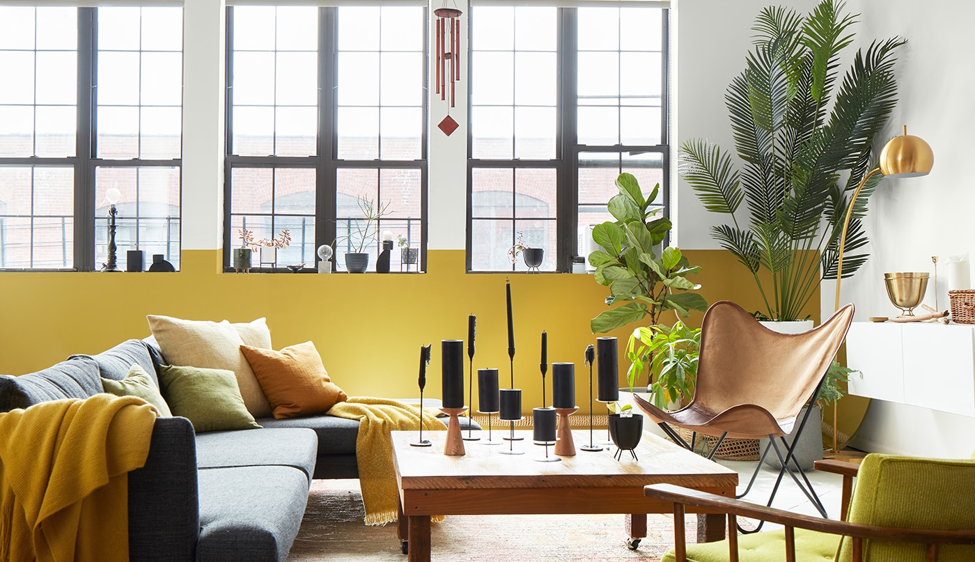 An airy, loft style living room with a white painted wall and ceiling with exposed pipes, a white and yellow accent wall with tall windows, and gray, yellow, and wood modern furnishings.