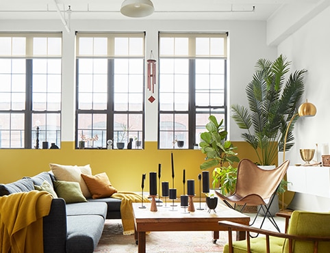 An airy, loft style living room with a white painted wall and ceiling with exposed pipes, a white and yellow accent wall with tall windows, and gray, yellow, and wood modern furnishings.