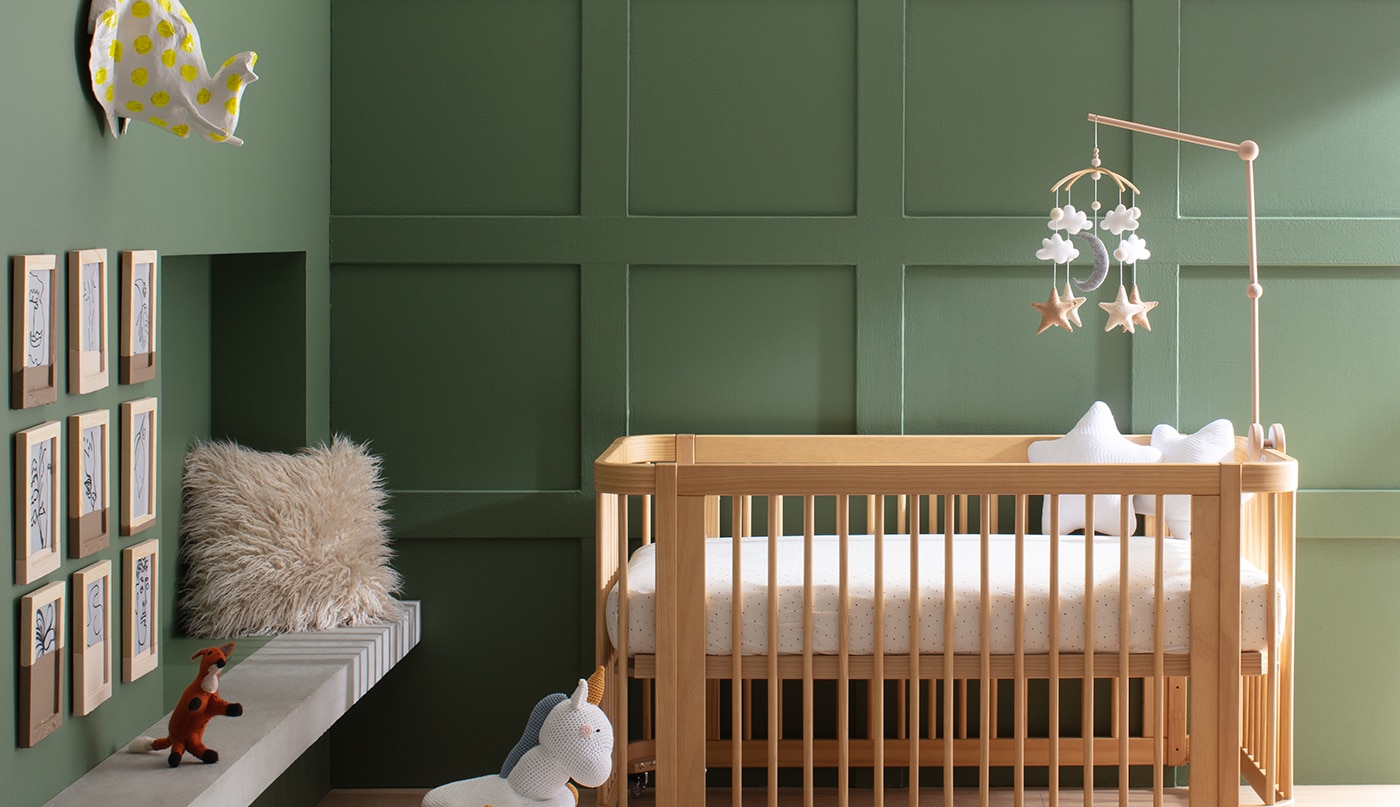 A sunlit baby room with a modern wood crib against a rich green-painted panelled wall, and adjacent green wall with colourful wall-mounted animal heads, framed artwork, and a multi-coloured rug.