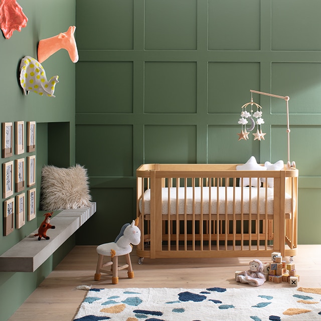 A sunlit baby room with a modern wood crib against a rich green-painted panelled wall, and adjacent green wall with colourful wall-mounted animal heads, framed artwork, and a multi-coloured rug.