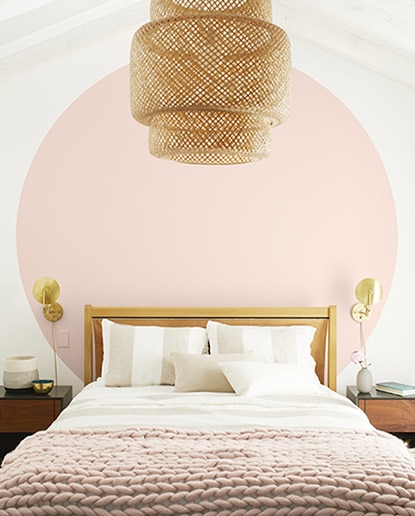 Light bedroom with white walls and a large light pink circle behind white bed with pink throw blanket and a basket chandelier.