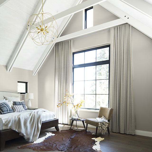 Chambre à coucher ensoleillée aux murs gris pâle et plafond cathédrale blanc avec poutres et grande fenêtre à carreaux.