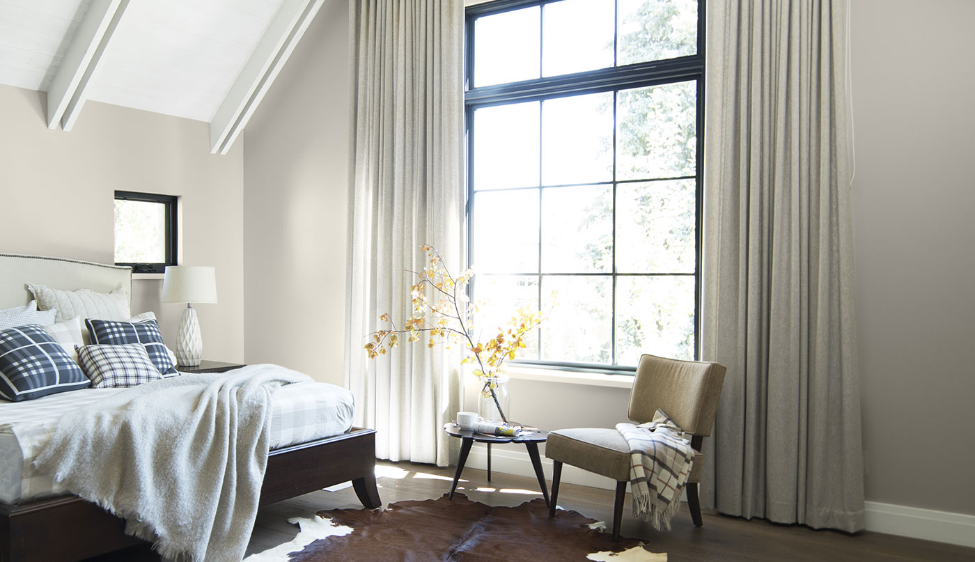 Chambre à coucher ensoleillée aux murs gris pâle et plafond cathédrale blanc avec poutres et grande fenêtre à carreaux.