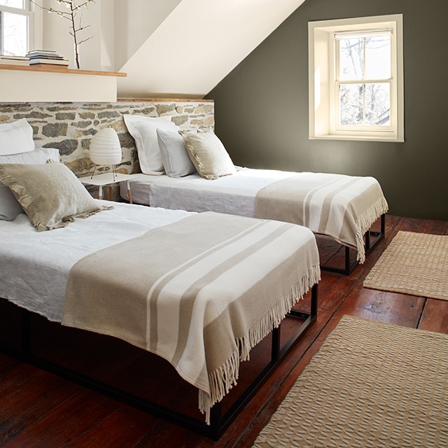 A guest room with an off-white-painted window trim set within a brownish-green painted accent wall, white-painted walls and ceiling, and a stone wall running the width of two beds with neutral bedding. 