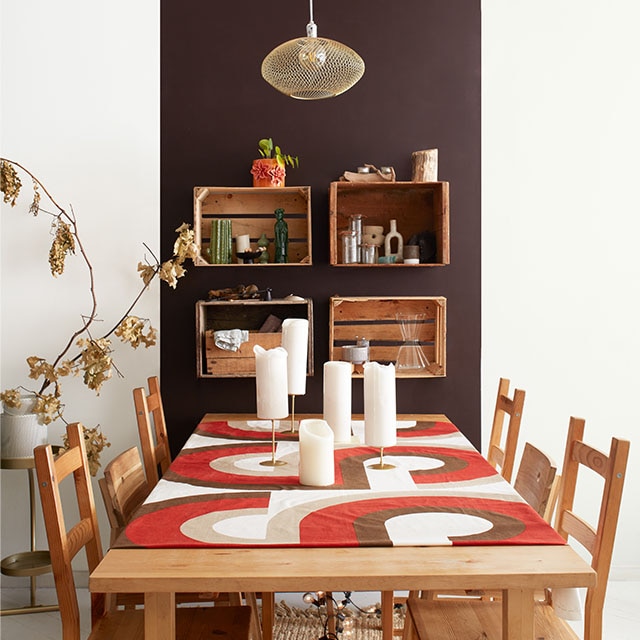 Salle à manger d’un appartement moderne avec panneau brun sur mur blanc, étagères murales créées à partir de palettes en bois, table et chaises en bois avec chemin de table orange et brun.