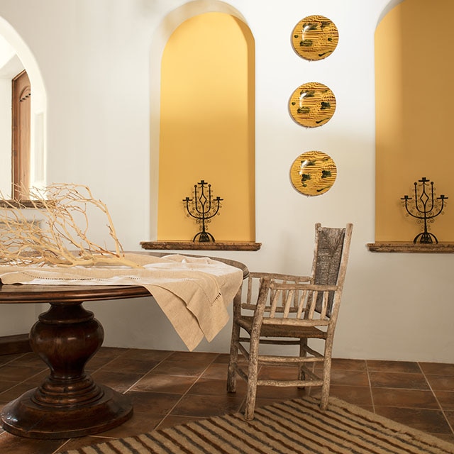 An airy, white-painted dining room with two yellow-painted alcoves, yellow decorative plates, dark wood ceiling beams, a rustic metal chandelier, and a round wood table and chair. 