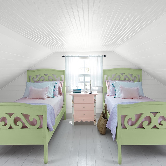 A cozy white-painted attic bedroom with shiplap walls and ceiling shows off two green twin beds with multi-colored pastel bedding, flanking a window and small pink chest of drawers.