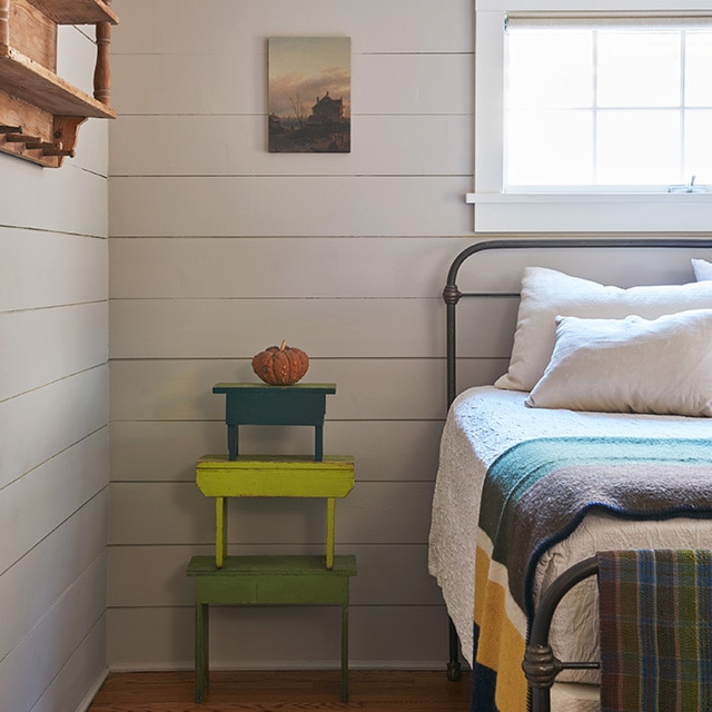 Coin d’une chambre douillette aux murs en planches à feuillure beiges avec lit en fer noir et couverture multicolore en dessous d’une fenêtre à boiseries blanches avec plusieurs bancs empilés dans différentes nuances de vert.