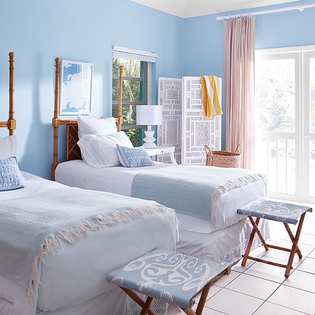 A bedroom with blue painted walls, a white ceiling and trim, twin beds and folding stools in blue and white decor.