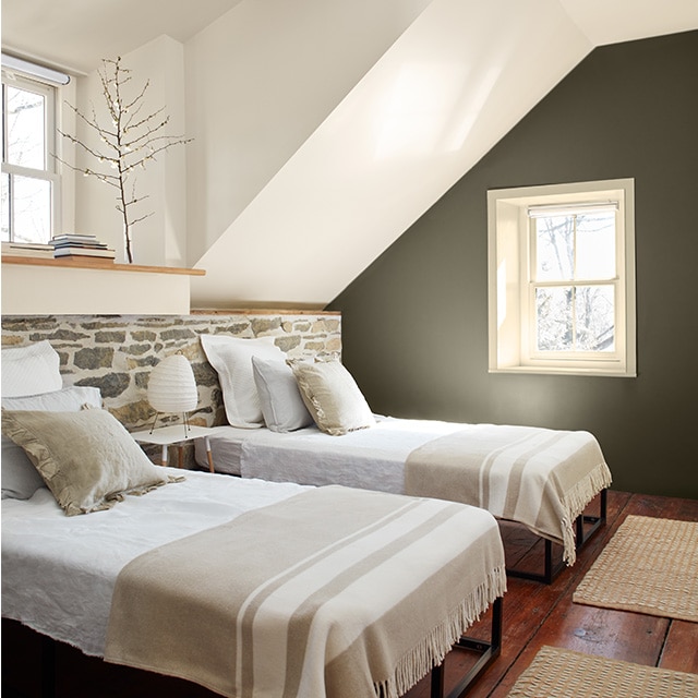 A cozy white-painted bedroom with a moss green accent wall, two twin beds in front of a low stone wall, hardwood floors and two small windows.