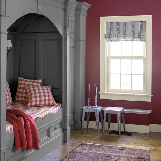 A gray-painted alcove bed with panelled inner walls, elegant moulding and red checkered bedding creates a cozy nook in a plum painted bedroom with a white trimmed window and ceiling.