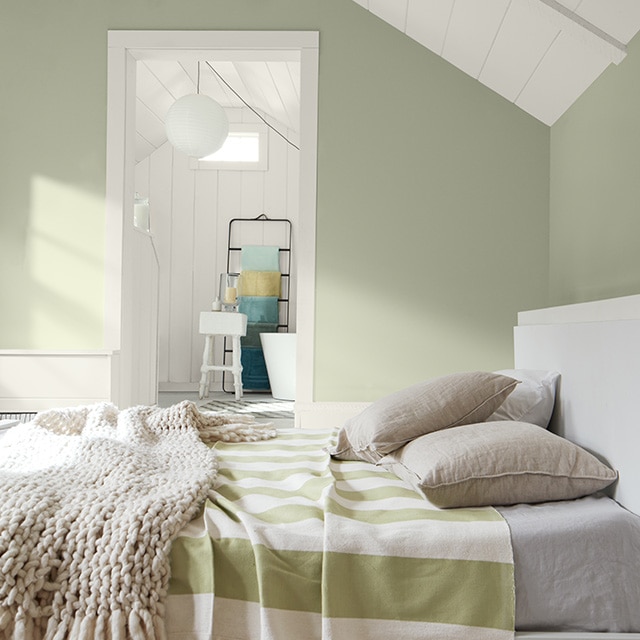 A sunlit sage green painted bedroom features a white ceiling and trim, a white bed with a green and white striped blanket, and an open door into a white painted en suite bathroom.