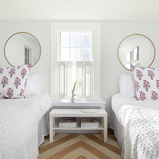 A white painted bedroom with two circular wall mirrors, twin beds with white bedding, a white shuttered window and striped jute rug.