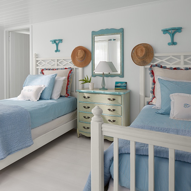 A cheerful, white-painted bedroom features two twin beds with white bed frames and blue bedding, flanking a pale yellow-green dresser with blue trim and a blue framed mirror.