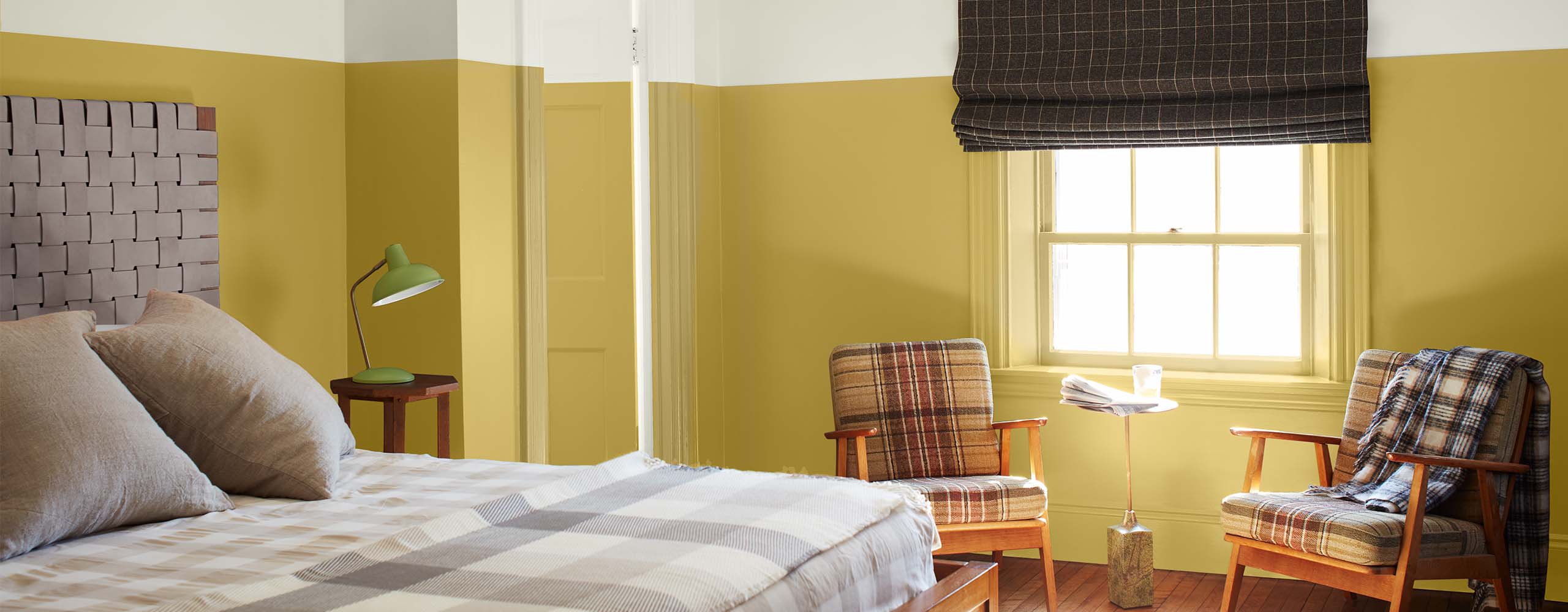 A bright two-tone painted bedroom with white-painted upper walls, upper door and ceiling, yellow-painted lower walls and door, tan checked bedding and two plaid chairs.