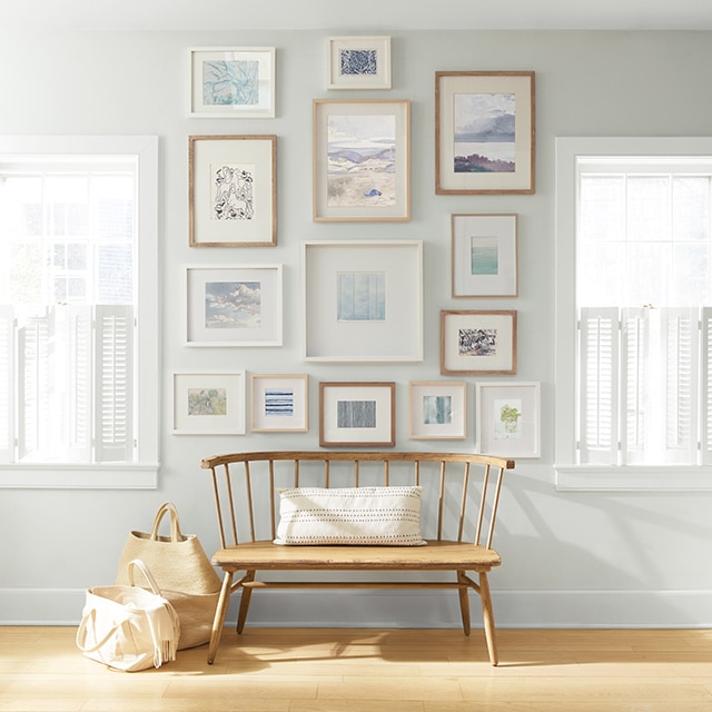 An off-white painted wall with a collection of framed art behind a wooden bench and shopping bags.