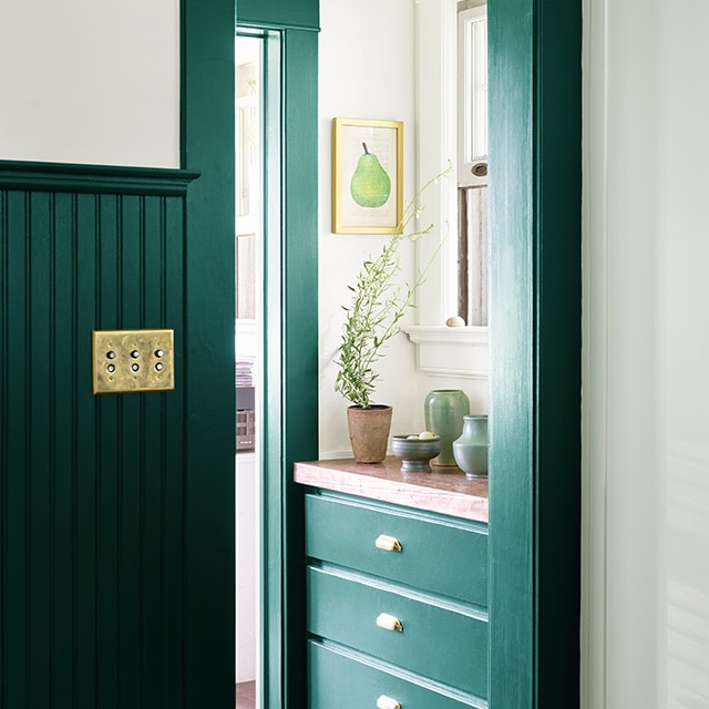 A hallway entrance with rich, dark green painted wainscotting, trim and built-in drawers underneath a window, and pale green painted wall cabinets.