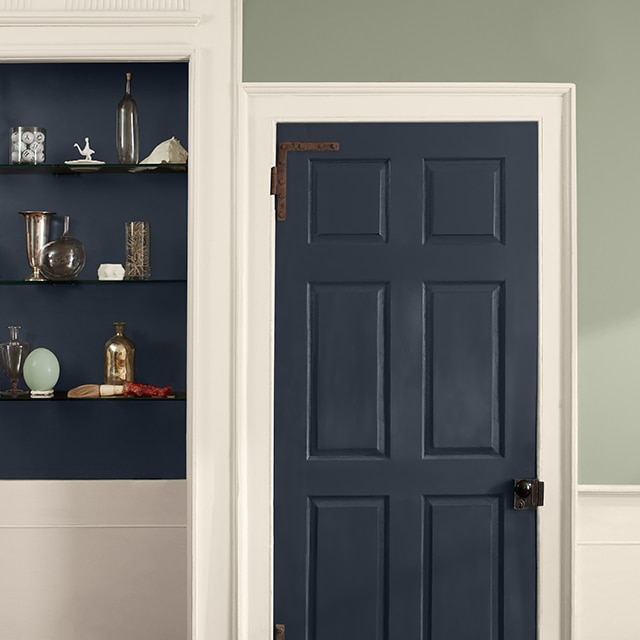 A two-tone wall painted light-sage green on the upper wall and off-white on the lower wall, with off-white trim and a dark blue door and inset shelves holding glass vases and trinkets.