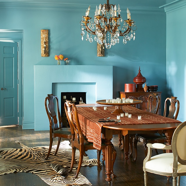 Une somptueuse salle à manger arborant des murs, un plafond et un foyer bleus, ainsi qu’un lustre suspendu au-dessus d’une grande table et de chaises en bois. Un style classique doté d’une touche d’exotisme.