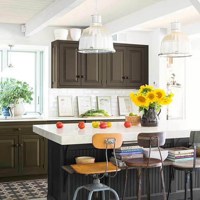 Une cuisine aérée de style industriel est agrémentée de couleurs foncées, avec la base noire de son îlot au comptoir blanc et ses armoires d’un vert-brun foncé.