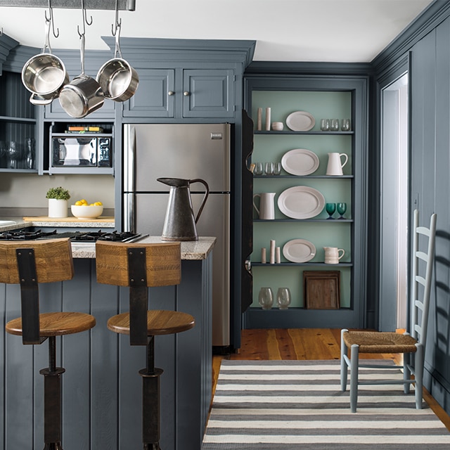A dark gray painted kitchen featuring beautiful built-in kitchen shelving with a light blue back wall and gray shelves holding white ceramic tableware and glassware, next to a stainless-steel refrigerator.