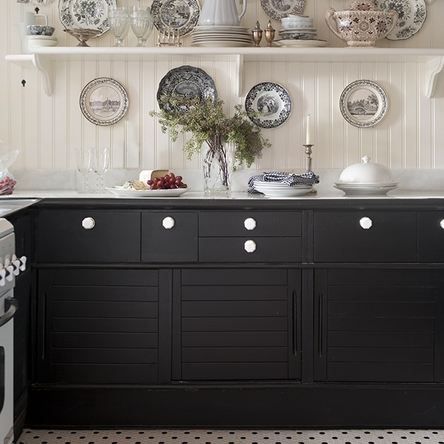 A white-painted French country kitchen with open shelving and patterned plates above black-painted cabinets, and a black and white tile floor.