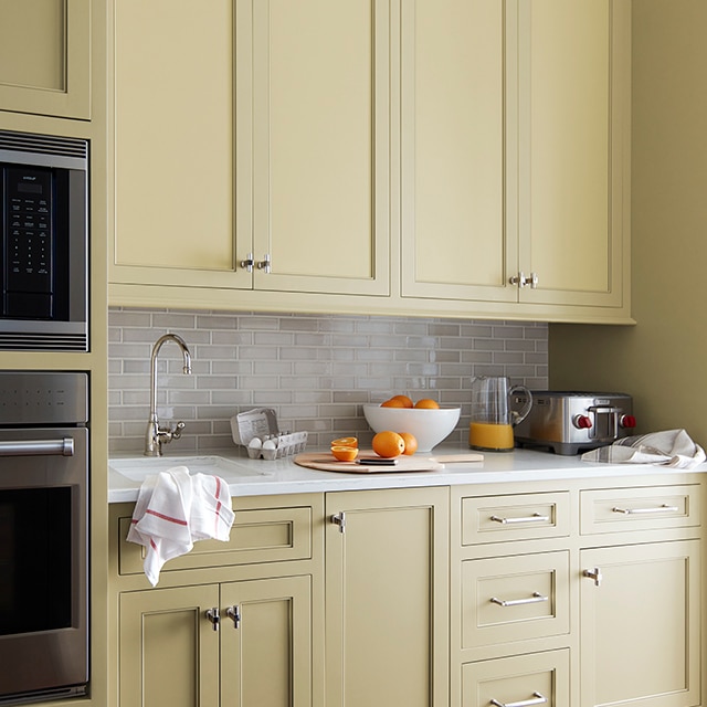 Gros plan de grandes armoires de cuisine enduites d’un beige doux aux reflets verts avec dosseret en carreaux blanc cassé et comptoir blanc avec quelques oranges.