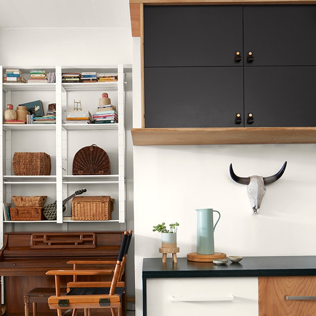 A small section of an open plan kitchen showcases cabinets and drawers in black and white paint with decorative shelving and a high director’s chair beyond.